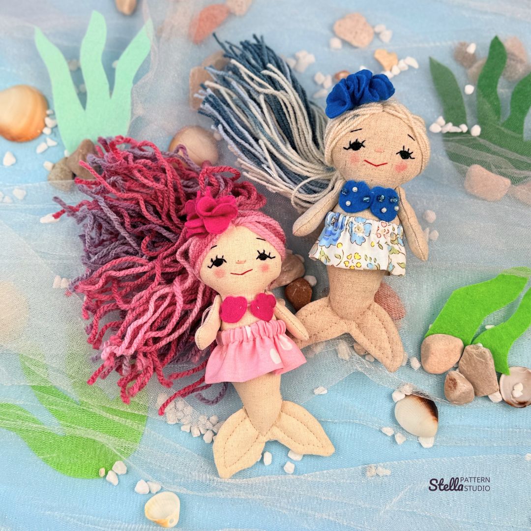 Two handmade dolls, one with pink hair and mermaid tail, the other with blonde hair and blue flower headband, on a blue fabric background with green leaves and shells.