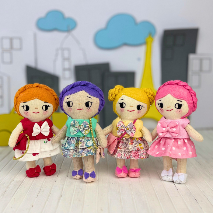 Four handmade fabric doll toys with colorful yarn hair and patterned dresses standing hand-in-hand on a light wood surface, with a stylized cityscape backdrop featuring blue clouds and a yellow Eiffel Tower silhouette.