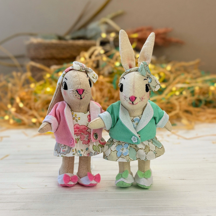 Two plush rabbit toys in dresses standing on a wooden surface with a neutral background.